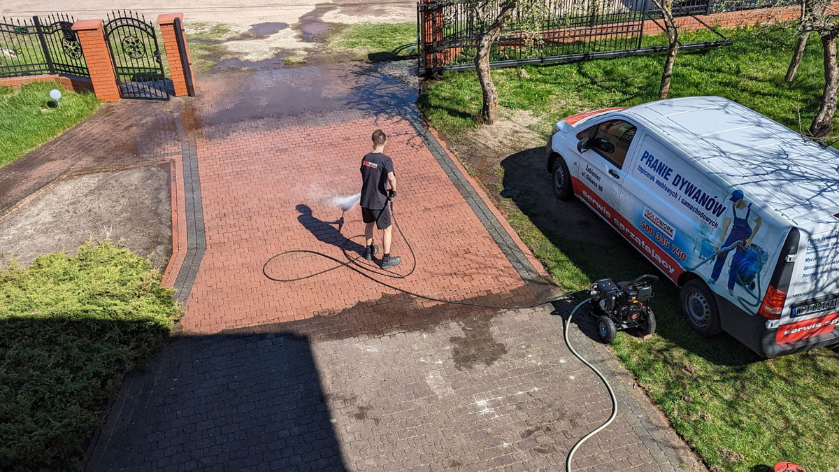 Czerwona kostka brukowa silnie zabrudzona w trakcie czyszczenia przez pracownika firmy Solid Work, specjalną maszyną, fragmenty już czyste wyglądają jak nowe. Obok zaparkowana furgonetka firmy oklejona jej reklamą.