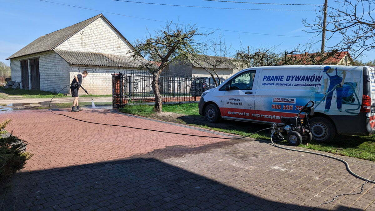 Czerwona kostka brukowa silnie zabrudzona w trakcie czyszczenia przez pracownika firmy Solid Work, specjalną maszyną, fragmenty już czyste wyglądają jak nowe. Obok zaparkowana furgonetka firmy oklejona jej reklamą.