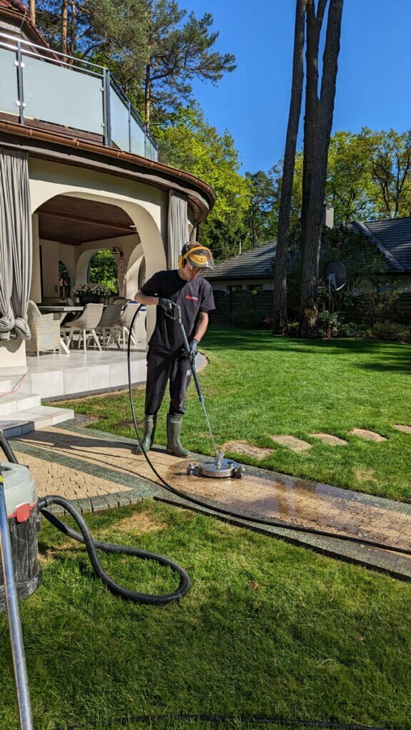 Mycie ścieżki z terakoty przecinającej trawnik przydomowy przez pracownika firmy Solid Work. Pracownik, zgodnie z zasadami bhp, ma twarz osłoniętą specjalną maską ochronną i jest w słuchawkach zabezpieczających przed hałasem