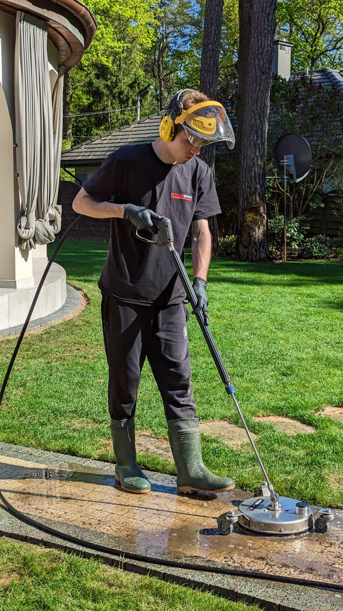 Mycie ścieżki z terakoty przecinającej trawnik przydomowy przez pracownika firmy Solid Work. Pracownik, zgodnie z zasadami bhp, ma twarz osłoniętą specjalną maską ochronną i jest w słuchawkach zabezpieczających przed hałasem