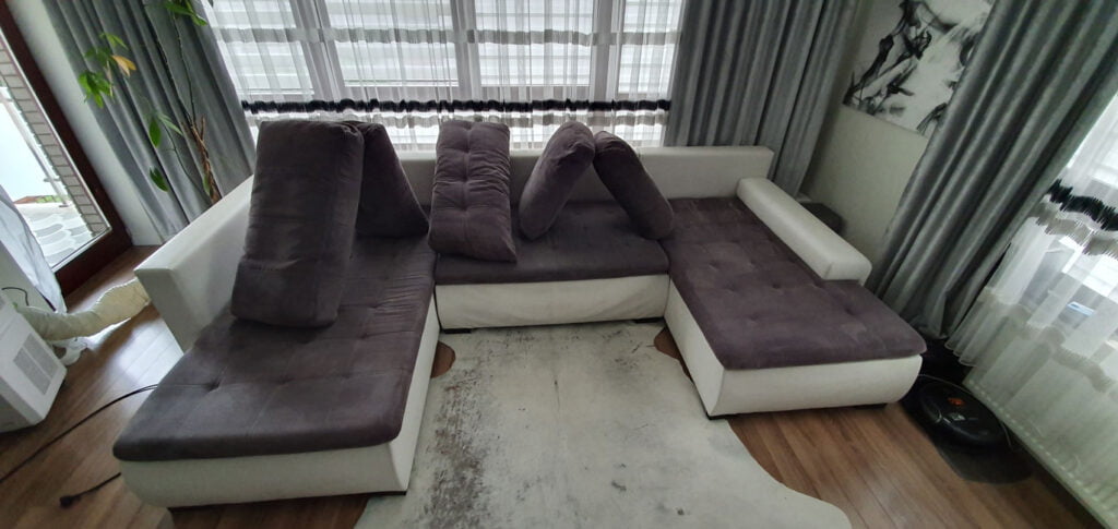 Nowoczesny wielki narożny komplet wypoczynkowy do salonu - trzy legowiska w kolorze jasnoszarym spody i brązowym na górze, oraz wielkie poduchy. Wszystko pięknie odświeżone