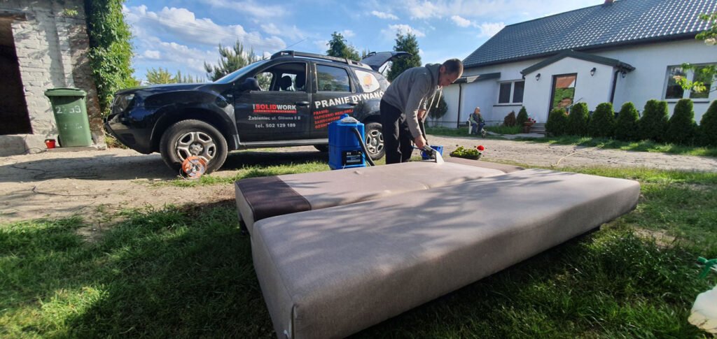 Odkurzanie leżącego na trawie siedzenia zdjętego z rozkładanej kanapy. W tle samochód firmowy Solid Work